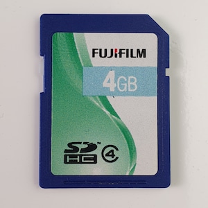 May include: A blue Fujifilm SDHC memory card with a 4GB storage capacity. The label features a green abstract wave design, the Fujifilm logo, the text 4GB on a light blue background, and the SDHC and class 4 speed rating symbols in black.
