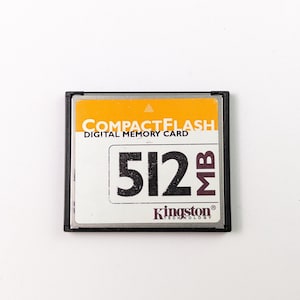 May include: A CompactFlash digital memory card with a black frame. The card has an orange top section with the words "COMPACTFLASH DIGITAL MEMORY CARD" in white. The card displays "512 MB" in large black numbers and the Kingston Technology logo.