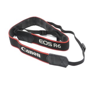 Può includere: Tracolla per fotocamera Canon EOS R6 nera con bordi rossi e scritte bianche. La tracolla ha una sezione imbottita per il collo e lunghezza regolabile. Il logo Canon e il nome del modello EOS R6 sono visibili.