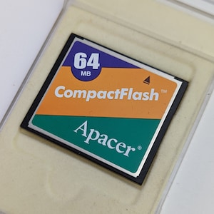 May include: A 64 MB CompactFlash memory card in a clear plastic case. The card has a black border, orange and green sections, and the "CompactFlash" and "Apacer" brand names are visible.