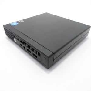 May include: A black HP desktop computer. The device has a rectangular shape with a flat top and a series of ports on the front. A blue sticker is visible on the top left corner. The computer is on a white surface.