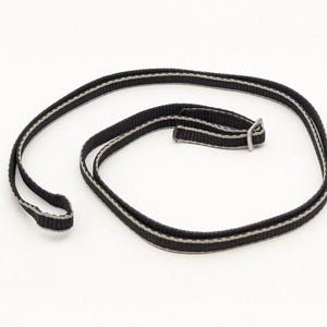 Pode incluir: Uma alça de nylon preta com finas listras cinzas ao longo de ambas as bordas. A alça está enrolada em formato circular e inclui uma pequena fivela deslizante retangular de metal prateado para ajuste de comprimento, criando um acessório funcional.