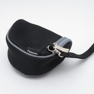 Pode incluir: Um estojo de transporte preto, semirrígido e curvo, com acabamento de zíper cinza e uma alça de pulso preta. O estojo apresenta uma pequena etiqueta preta com o texto branco Panasonic costurado perto do puxador de metal prateado.