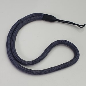 Pode incluir: Uma alça de pulso de cordão trançado cinza-escuro com um conector de plástico preto liso e um laço fino de nylon preto na extremidade. O cordão tem uma superfície trançada texturizada e está disposto em uma forma curva e flexível.
