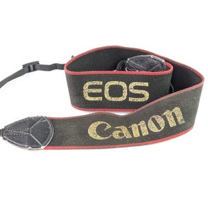 Pode incluir: Uma alça de pescoço preta para câmara com o texto EOS e Canon em letras douradas. A alça é feita de tecido com debrum vermelho nas bordas e inclui pontos de fixação em material semelhante a couro preto em cada extremidade.