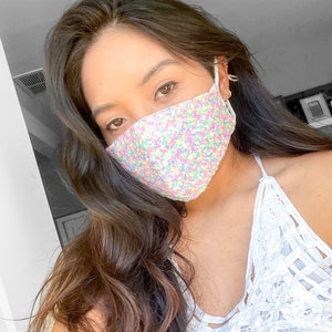 May include: A woman wearing a white lace top with a sequined face mask. The mask is white with multi-colored sequins.