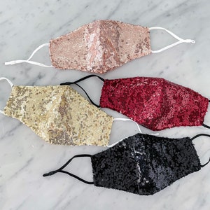 May include: Four face masks made of sparkly sequins. The masks are in gold, black, pink, and red.
