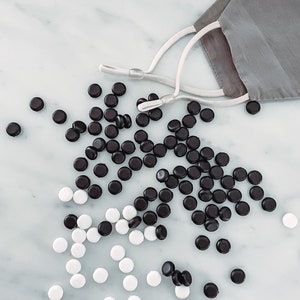 May include: A close-up of a pile of black and white round buttons. The buttons are scattered on a white marble surface. A gray face mask with white elastic straps is lying on the surface.