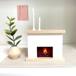 Puede incluir: Chimenea blanca en miniatura con una pantalla de fuego falso, coronada con dos candelabros blancos. Un pequeño jarrón de madera con hojas verdes está a la izquierda. Un tapiz de pared rosa está en el fondo. La chimenea es de madera.