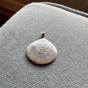 Puede incluir: Un colgante de plata en forma de lágrima con acabado mate y un motivo floral grabado en el centro. La flor tiene un núcleo circular dorado con pequeñas piedras incrustadas y está rodeada por diminutos puntos dorados y pétalos grabados.