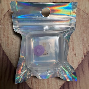 May include: A clear, square plastic case with a purple object inside, sealed in a holographic, iridescent pouch. The pouch has a small hole at the top and is sealed shut. The background is a light brown wooden surface.