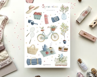 May include: A sheet of stickers with a spring theme. The stickers feature a bicycle, a picnic basket, a thermos, a camera, a book, a hat, sunglasses, a lemon tree, a bird, and a picnic blanket. The stickers are on a white background and have a light blue and pink color scheme. The text "Spring Vibes" is at the top of the sheet.