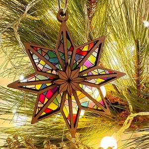 May include: A wooden star ornament with a stained glass design in shades of red, blue, green, yellow, and purple. The star is hanging from a pine tree branch with white lights.
