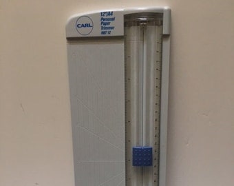 May include: A gray paper trimmer with a clear ruler guide and a blue locking mechanism. The trimmer has a cutting length of 12 inches and features the brand name "CARL". The trimmer is designed for personal use and is suitable for trimming paper.