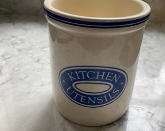 May include: White ceramic utensil holder with a blue and white stripe around the top and a blue oval with the words "Kitchen Utensils" in white.