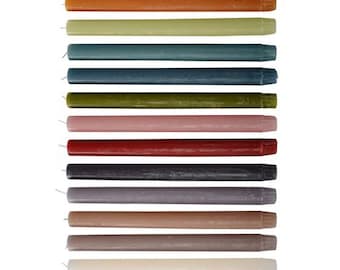 Op de afbeelding: Assortiment van 14 tapers in verschillende kleuren, waaronder bruin, oranje, groen, blauw, roze, rood, grijs, beige en wit. De kaarsen zijn verticaal in een rij gerangschikt.