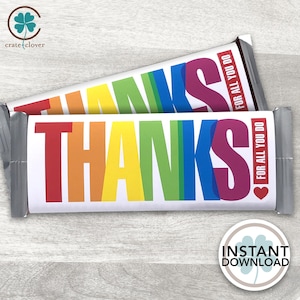 Puede incluir: Dos envoltorios de barras de chocolate con la palabra «THANKS» en colores arcoíris. El texto «FOR ALL YOU DO» está impreso en rojo en el lateral. Un símbolo de corazón está al lado del texto. Los envoltorios están sobre una superficie de madera clara.