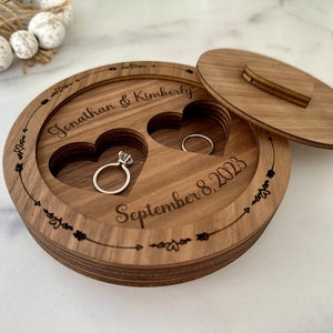 May include: A round wooden ring box with two heart-shaped ring slots. The box is engraved with the names "Jonathan & Kimberly" and the date "September 8, 2023". The box has a lid and is decorated with arrows and floral accents.