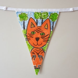 May include: A hand-drawn triangular bunting flag with a cartoon tiger design. The tiger is orange with black stripes and green eyes, set against a backdrop of green trees and a blue sky. The flag is attached to a white ribbon.