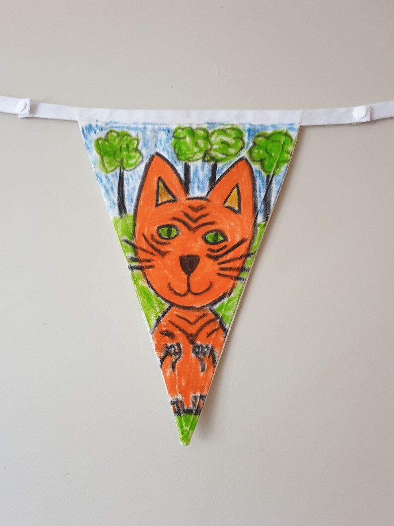 May include: A hand-drawn triangular bunting flag with a cartoon tiger design. The tiger is orange with black stripes and green eyes, set against a backdrop of green trees and a blue sky. The flag is attached to a white ribbon.