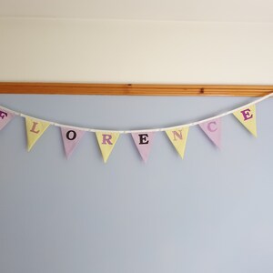 May include: A pastel-coloured fabric banner with the name "FLORENCE" spelled out in purple letters. The banner is strung up on a wall.