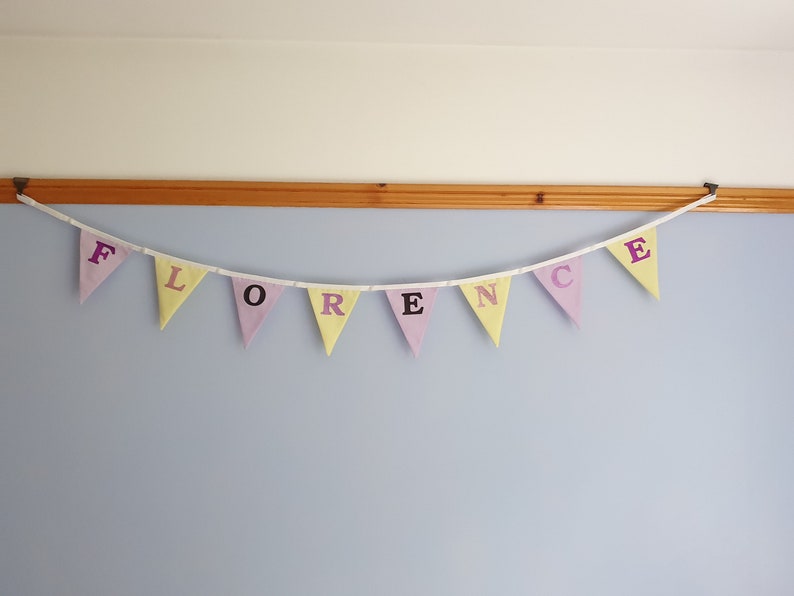 May include: A pastel-coloured fabric banner with the name "FLORENCE" spelled out in purple letters. The banner is strung up on a wall.