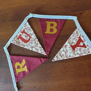 May include: A red and white fabric banner with the word "RUBY" spelled out in gold and red glitter letters. The banner is made of a white fabric with a red floral pattern and has blue ribbon ties.