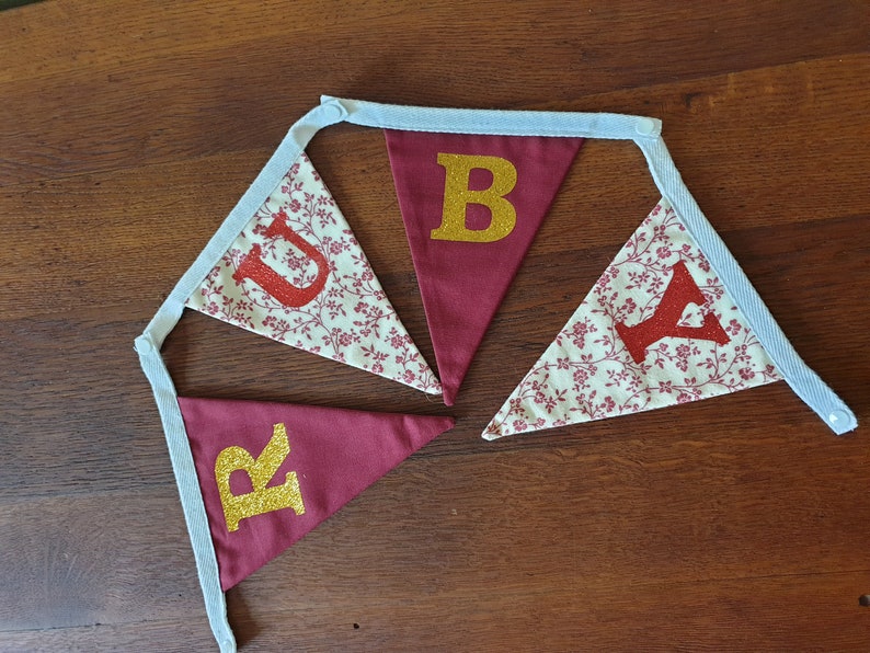 May include: A red and white fabric banner with the word "RUBY" spelled out in gold and red glitter letters. The banner is made of a white fabric with a red floral pattern and has blue ribbon ties.