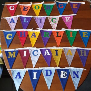 May include: A colourful bunting with the names George, Alice, Charlie, Micah, and Aiden spelled out in felt letters. Each letter is on a separate triangular flag.
