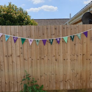 May include: A colourful banner with the text "Donna is 70" hanging on a wooden fence. The banner is made of triangular flags with different colours and patterns.