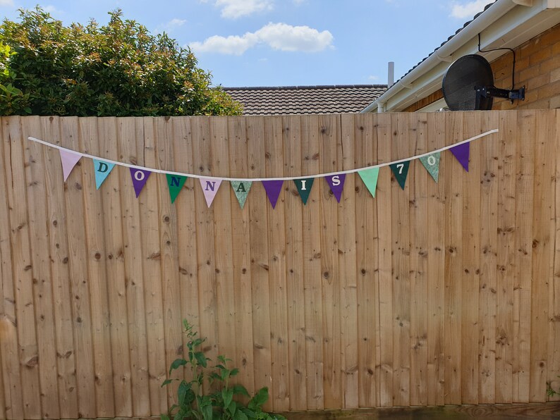 May include: A colourful banner with the text "Donna is 70" hanging on a wooden fence. The banner is made of triangular flags with different colours and patterns.