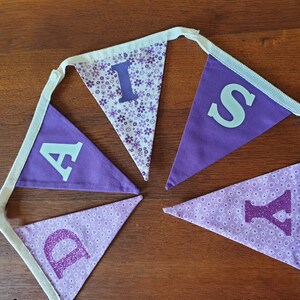 May include: Purple and white fabric banner with the word "Daisy" spelled out in white and purple letters. The letters are decorated with glitter and small purple flowers.