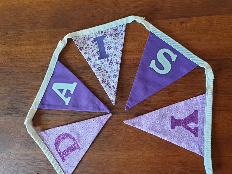 May include: Purple and white fabric banner with the word "Daisy" spelled out in white and purple letters. The letters are decorated with glitter and small purple flowers.