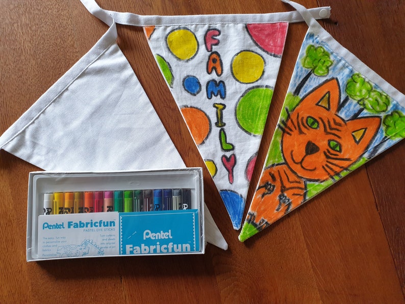 May include: A collection of white fabric bunting, one plain, two decorated with colourful drawings and the word "FAMILY". A box of Pentel Fabricfun pastel dye sticks is also included, ideal for creating custom designs on the bunting.