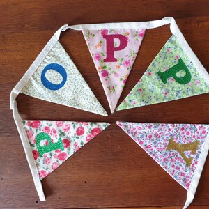 May include: A bunting banner with five flags, each featuring a different letter of the word "HOPE" in glitter. The flags are made of white fabric with a floral pattern and are connected by white ribbon.