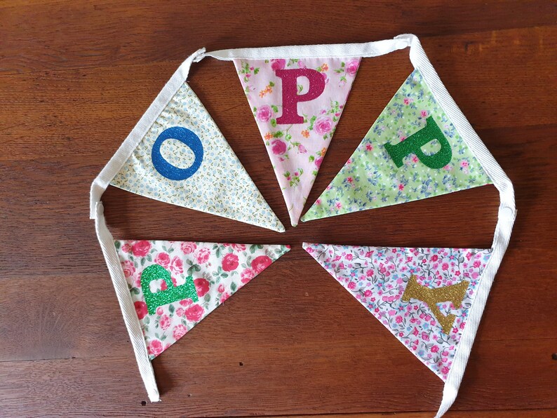 May include: A bunting banner with five flags, each featuring a different letter of the word "HOPE" in glitter. The flags are made of white fabric with a floral pattern and are connected by white ribbon.