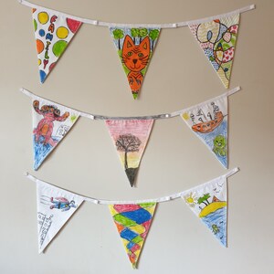 May include: A collection of triangular fabric bunting flags, each hand-drawn with unique designs. The illustrations include a cat, a tree, and abstract patterns. The flags are strung on white ribbon and displayed against a plain wall.
