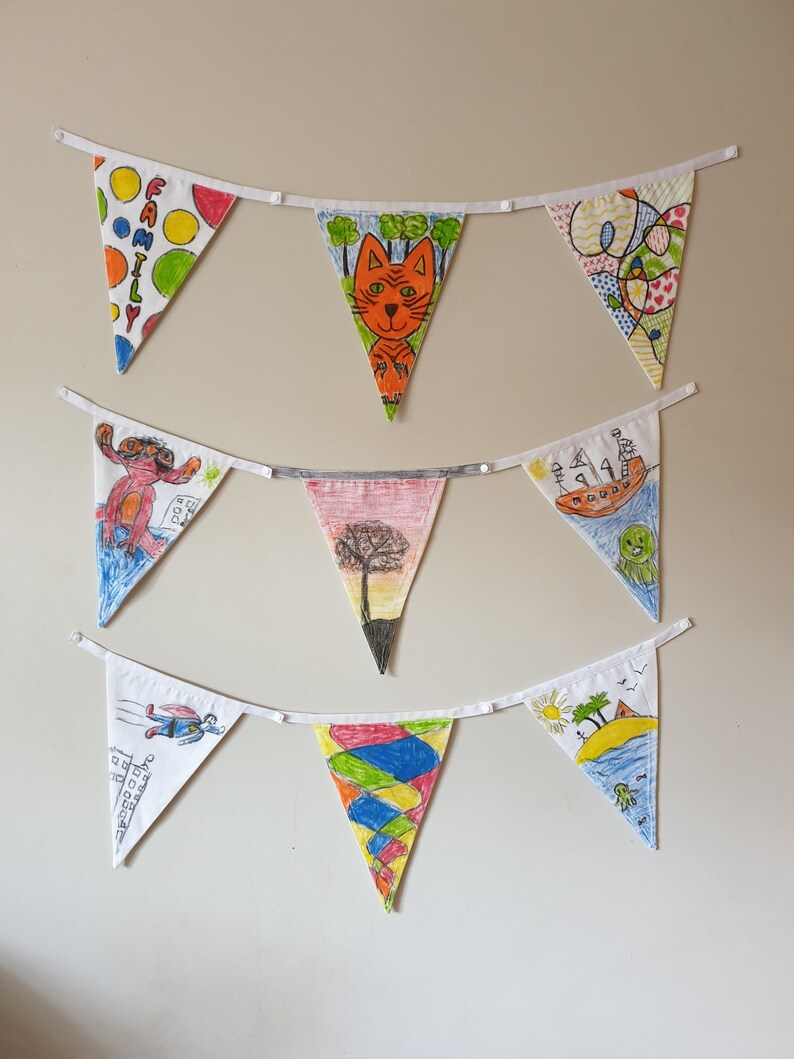 May include: A collection of triangular fabric bunting flags, each hand-drawn with unique designs. The illustrations include a cat, a tree, and abstract patterns. The flags are strung on white ribbon and displayed against a plain wall.