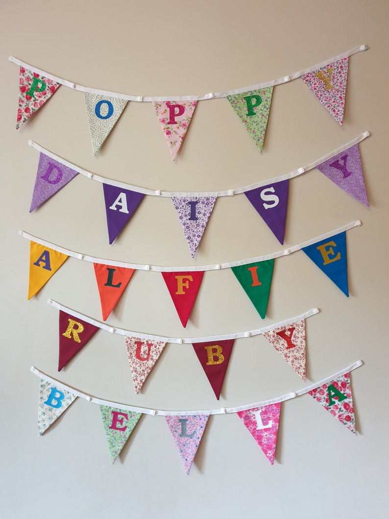 May include: A colourful fabric banner with the names Poppy, Daisy, Alfie, Ruby, and Bella spelled out in individual triangular flags. Each flag has a different colour and pattern.