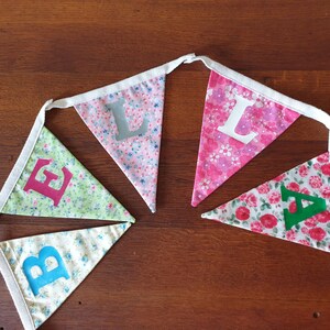 May include: A bunting with five flags, each with a different floral pattern and a letter on it. The letters spell out "BELLA".