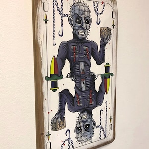 May include: A wooden playing card with a white background and a graphic of a character from the horror movie Hellraiser. The character is depicted with a hooked chain, a box, and a menacing expression. The card is labeled with the letter "J" for Jack.