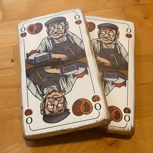 May include: Two wooden playing cards featuring a cartoon illustration of a man in a cap and apron, with a mustache and glasses. The cards have a distressed, aged look, with brown and white tones. Each card has a pretzel and the number zero.