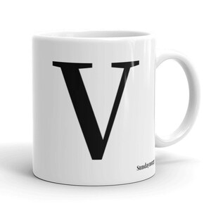Pode incluir: Caneca de cerâmica branca com uma letra V preta impressa na frente. A caneca tem uma alça do lado direito.