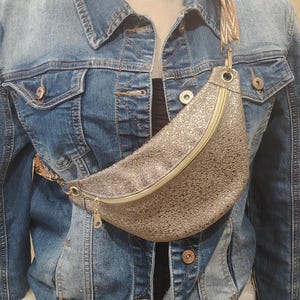 Op de afbeelding: Een spijkerjasje met een lichte wassing, gedragen over een zwarte top en een wit, pluizig kledingstuk. Een sprankelende, goudkleurige heuptas met ritssluiting en patroonband wordt over de borst gedragen. De heuptas is ongeveer 30 cm breed.