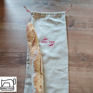 Könnte beinhalten: Eine weiße Stofftasche mit roten Kordelzugbändern und dem in Rot gestickten Text "Mon Pain". Ein langes Baguette ist teilweise in der Tasche.