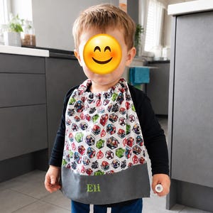 May include: A bib featuring a white background with a repeating pattern of superhero characters in hexagonal frames and a solid gray bottom panel. The name Eli is embroidered in green thread on the gray section. The bib has a gathered neckline.