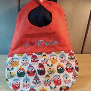May include: A red baby bib with a white button and a pocket. The pocket is lined with a fabric featuring a pattern of owls wearing winter hats and scarves. The bib is embroidered with the text "I love Mamie".