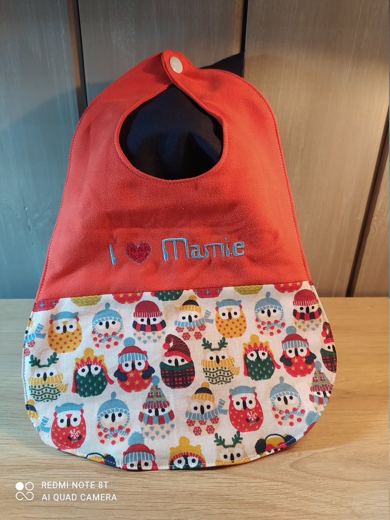 May include: A red baby bib with a white button and a pocket. The pocket is lined with a fabric featuring a pattern of owls wearing winter hats and scarves. The bib is embroidered with the text "I love Mamie".