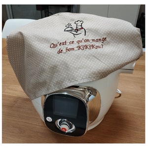 May include: A beige kitchen towel with white polka dots and embroidered text that reads "Qui'est ce qu'on mange de bon KikiKOU?"  The towel is draped over a white and silver kitchen appliance with a digital display.