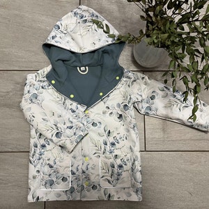 May include: A children's hooded jacket with a botanical print of grey and blue leaves on a white base. It has a teal lining, yellow buttons, and two front pockets. The jacket is designed for children.
